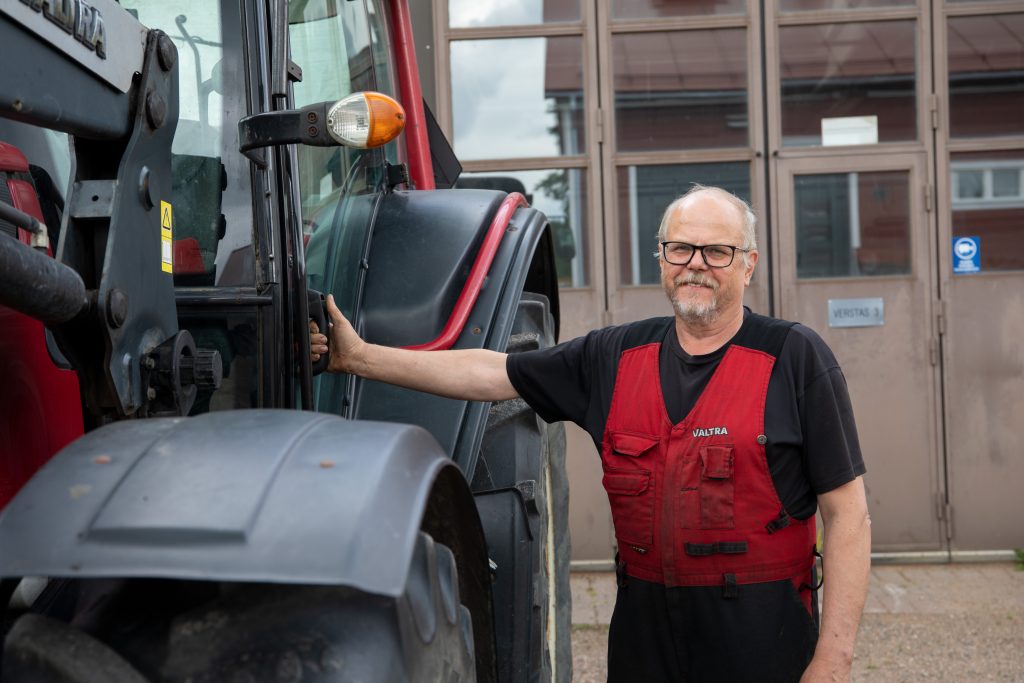 Henkilö seisoo traktorin vierellä pitäen traktorin ovesta kiinni. Hänellä on yllään työhaalarit. Taustalla näkyy suuri hallin ovi.