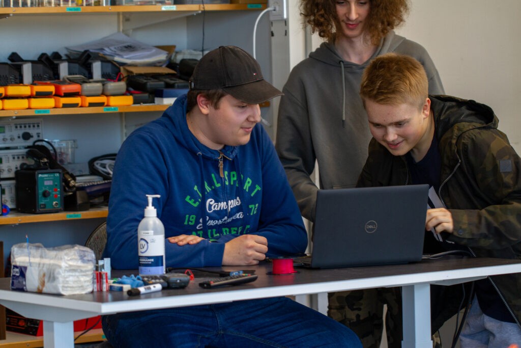 Kaksi henkilöä istuu teräksisen pöydän ääressä ja katsovat kannettavaa tietokonetta. Taustalla seisoo kolmas henkilö. Taustalla näkyy myös hylly, jonne on asetettu erilaisia elektroniikkatarvikkeita.