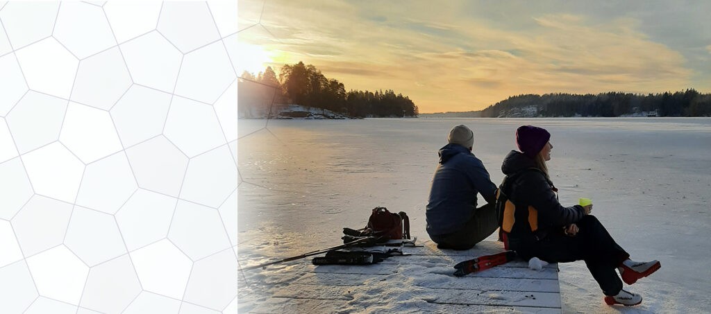 kaksi henkilöä istuu laiturin päässä jäisen järven rannalla katsoen auringon laskua