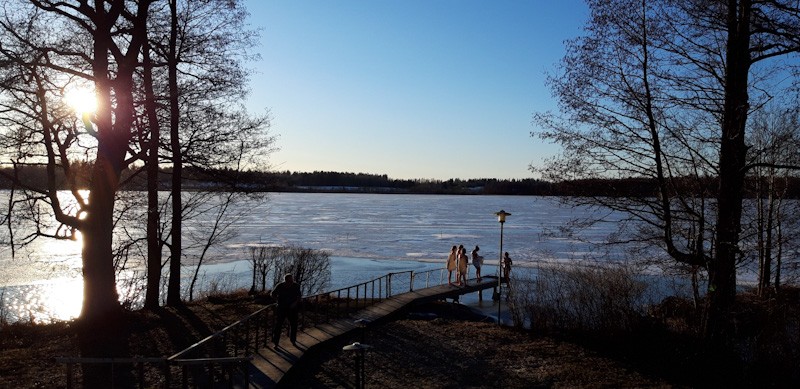 Keväinen järvimaisema, jossa uimareita laiturilla.